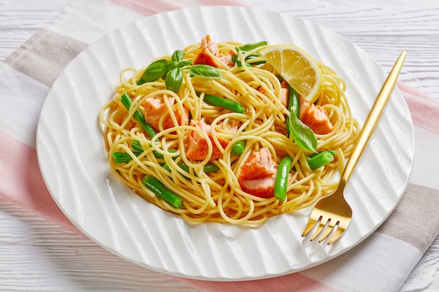 Délicieuses pâtes au saumon avec haricots verts et sauce au beurre à l'ail avec basilic frais sur le dessus et citron servi sur une assiette blanche avec une fourchette sur une table en bois blanc avec une serviette rose, gros plan