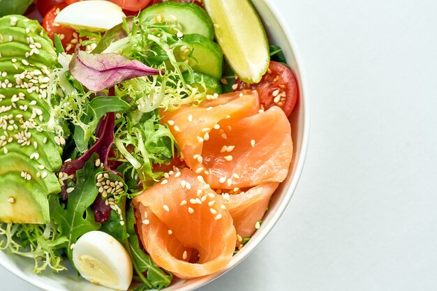 Délicieux bol de légumes avec saumon, avocat, concombre, tomates cerises et salade mixte dans une assiette blanche. Isolé sur une surface grise.