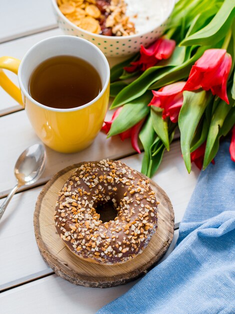 Délicieux Bol De Yaourt Avec Des Flocons De Maïs, Des Noix Et De La Confiture Sur Une Table En Bois Blanc. Concept De Nutrition Saine Et Biologique. Tulipes Avec Tasse De Thé, Beignet Et Petit-déjeuner. Vue De Dessus