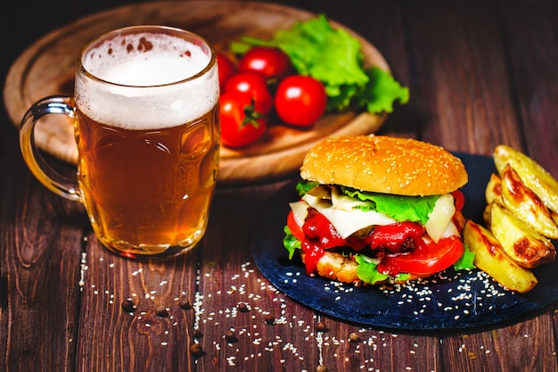 Un délicieux burger de bœuf avec de la laitue et des pommes de terre, un verre de bière servi sur une planche à découper en pierre. Sombre