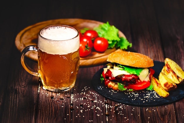 Un délicieux burger de bœuf avec de la laitue et des pommes de terre, un verre de bière servi sur une planche à découper en pierre. Sombre