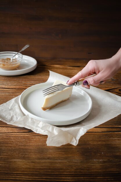 Délicieux cheesecake classique sucré dessert New York sur fond rustique en bois. Mains de femme coupant une tranche de délicieux gâteau sur une assiette en céramique avec une fourchette à dessert avec de la confiture de pommes.