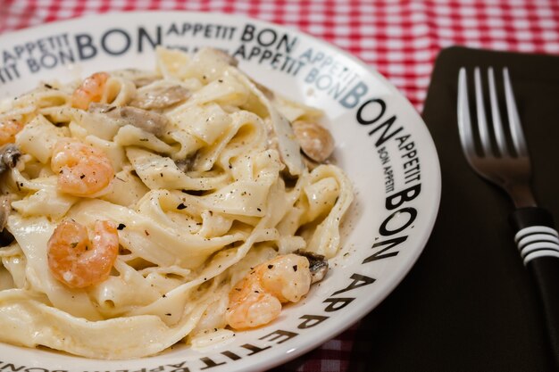 Délicieux fettuccine à la crème de champignons et crevettes sur une assiette qui dit Bon Appétit avec nappe à carreaux rouges. Cuisine italienne.