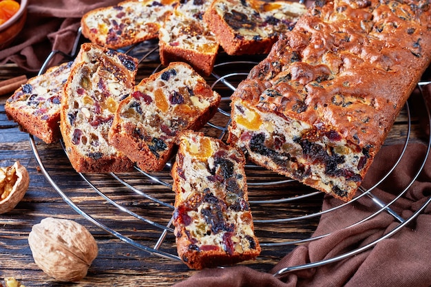 Délicieux gâteau riche en gros fruits secs sur un support de gâteau en fil avec un chiffon brun, des bâtons de cannelle, des abricots secs et des fruits de date sur une table en bois rustique, vue d'en haut, gros plan