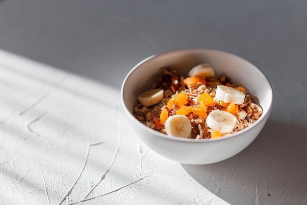 Délicieux muesli de petit-déjeuner sain avec de la farine d'avoine naturelle, des abricots secs, des noix et des bananes dans un bol blanc sur une table blanche au soleil le matin