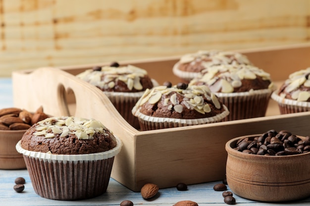 De délicieux muffins au chocolat sucrés, avec des pétales d'amandes dans un plateau en bois à côté de grains de café sur une table en bois bleue.