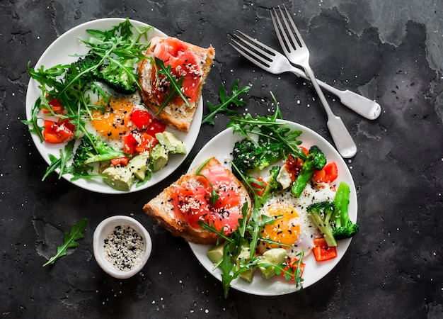 Délicieux petit-déjeuner sain oeuf avec salade de roquette aux légumes et pain grillé au fromage cottage au saumon sur fond sombre vue de dessus