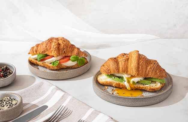 Délicieux petit-déjeuner avec sandwichs croissants à l'œuf poché, avocat, fromage à pâte molle, mozzarella et tomate dans une assiette sur fond blanc