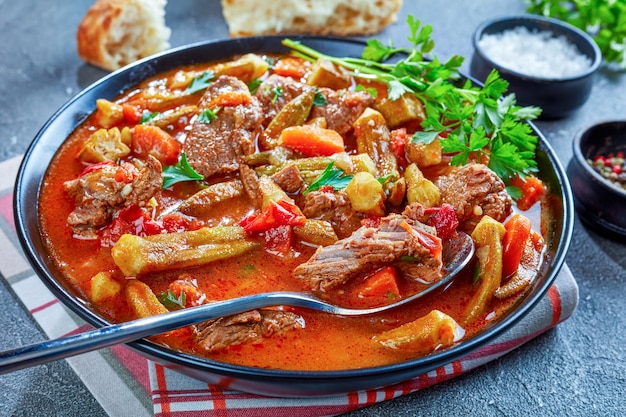 Délicieux ragoût de boeuf et de gombo ou soupe servi dans un bol avec cuillère, sur une table en béton gris avec du pain ciabatta, vue d'en haut, gros plan