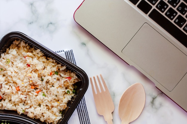 Délicieux riz frit dans la boîte à lunch sur la vaisselle avec une fourchette et une fourchette en bois et un ordinateur portable sur le lieu de travail