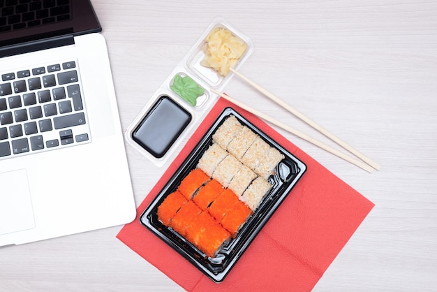 Délicieux rouleaux japonais au saumon, avocat, concombre, fromage, dans des emballages à emporter avec bâtonnets de bois, sauce soja et ordinateur portable sur la table de bureau Mise au point sélective Concept de nourriture de livraison asiatique