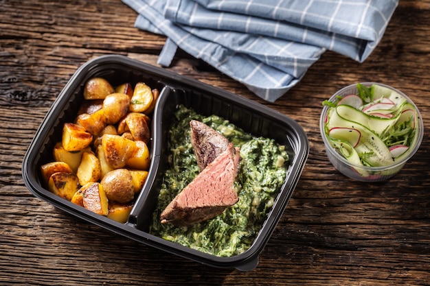 Délicieux steak de viande rouge sous vide emballé dans une boîte en plastique portable à emporter et servi avec des pommes de terre rôties croustillantes et une savoureuse trempette aux épinards et une salade de légumes frais.