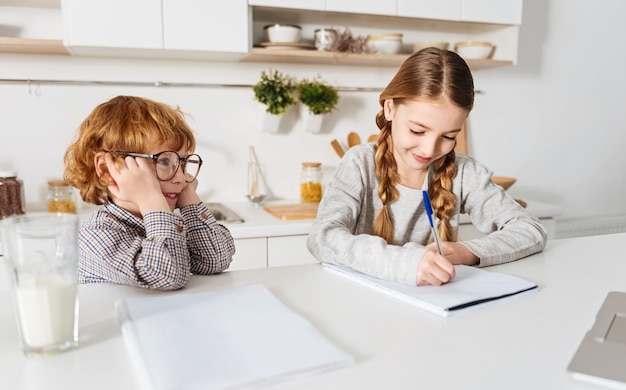 Démarrage actif. Jolie fille positive concentrée écrivant son devoir à la maison et son frère de litière la regardant pendant qu'ils passent tous les deux du temps dans la cuisine