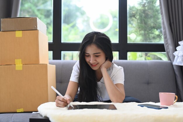 Démarrez une femme entrepreneur de petite entreprise travaillant avec une tablette numérique et assise entourée de boîtes à la maison.