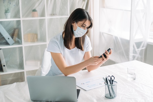 Déménagement de bureau Fermeture d'entreprise Chômage pandémique Femme perturbée portant un masque facial à la recherche d'un nouveau lieu de travail contactant un agent immobilier à l'aide d'un ordinateur portable dans une pièce lumineuse avec des meubles couverts