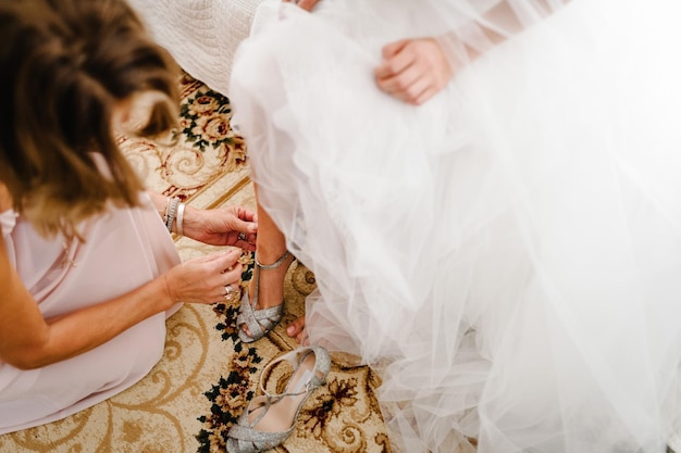 Demoiselles d'honneur mettre sur la jambe mariée élégantes chaussures argentées classiques Femme élégante mettant les mains sur des chaussures sur une robe blanche de fond Préparation du matin de mariage