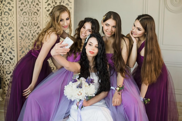 Les demoiselles d'honneur prennent un selfie sur un smartphone et rient le matin de la mariée à la maison De belles filles en robes de mariée violettes et une mariée avec un bouquet de pivoines s'amusent