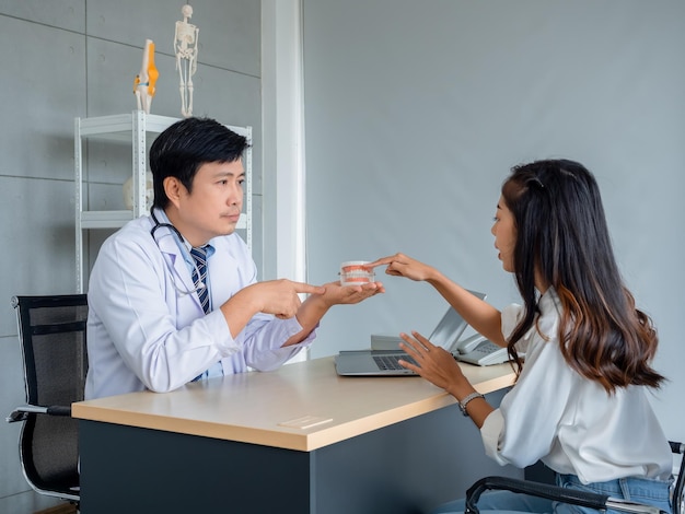 Dentiste adulte asiatique professionnel masculin tenant une prothèse dentaire modèle tout en expliquant et en éduquant une jeune belle patiente sur le bureau dans le bureau de la clinique médicale Concept dentaire oral