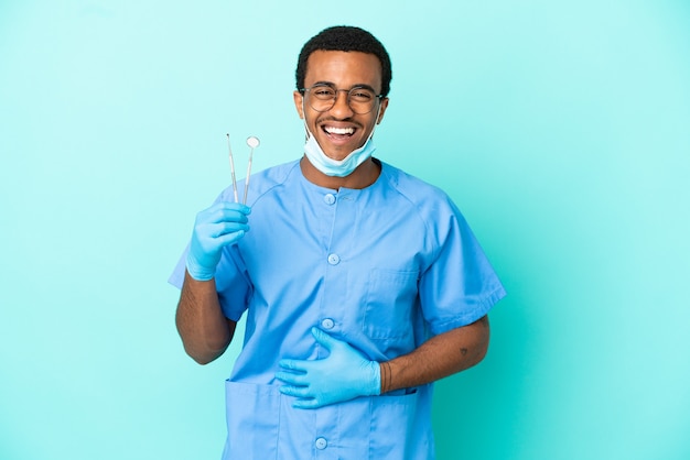 Dentiste afro-américain tenant des outils sur fond bleu isolé souriant beaucoup