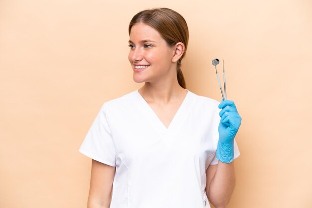 Dentiste caucasien femme tenant des outils isolés sur fond beige regardant sur le côté et souriant