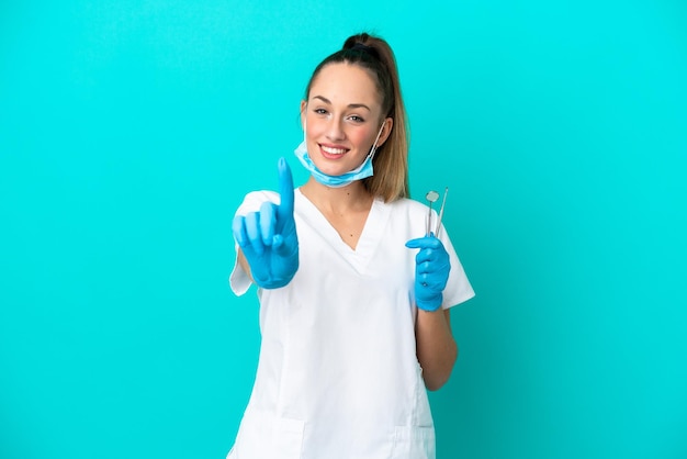 Dentiste caucasien femme tenant des outils isolés sur fond bleu montrant et levant un doigt