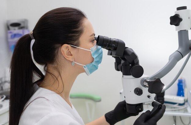 Une dentiste caucasienne aux cheveux noirs dans un masque de protection médicale travaille avec un microscope moderne dans une clinique dentaire