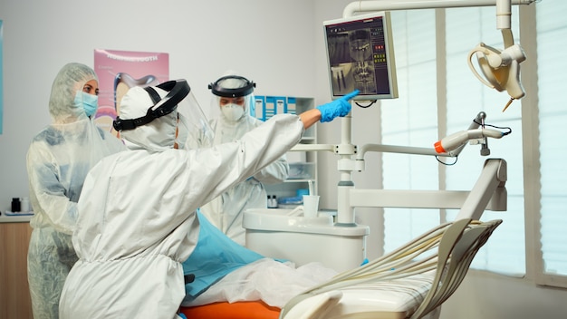 Dentiste en costume ppe pointant sur un moniteur dentaire numérique expliquant la radiographie à la mère et à l'enfant portant une combinaison pendant la pandémie mondiale. Nouvelle visite normale chez le dentiste lors d'une épidémie de coronavirus avec écran facial