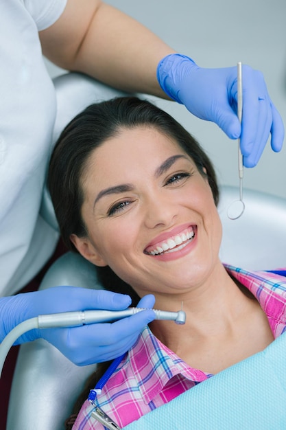 Dentiste dans des gants en caoutchouc bleu tenant une pièce à main à grande vitesse et un miroir buccal près du visage d'une femme souriante positive