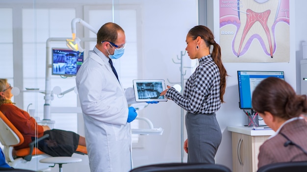 Dentiste dans la salle d'attente du cabinet dentaire parlant avec une patiente examinant une image radiographique sur une tablette pendant que les patients sont assis sur des chaises dans la zone de réception. Docteur montrant la radiographie dentaire, gadget moderne