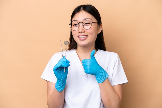Dentiste femme chinoise tenant des outils isolés sur fond beige donnant un geste du pouce levé