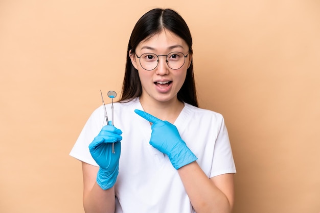 Dentiste femme chinoise tenant des outils isolés sur fond beige avec une expression faciale surprise