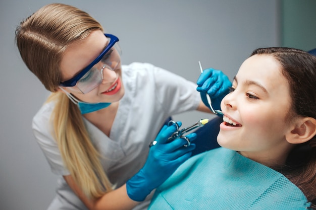 Dentiste femme prudente tenir les outils près de la bouche de la jeune fille. L'enfant montre les dents de devant. Elle est assise calmement dans un fauteuil dentaire.