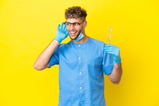 Dentiste homme blond tenant des outils isolés sur fond écoutant quelque chose en mettant la main sur l'oreille