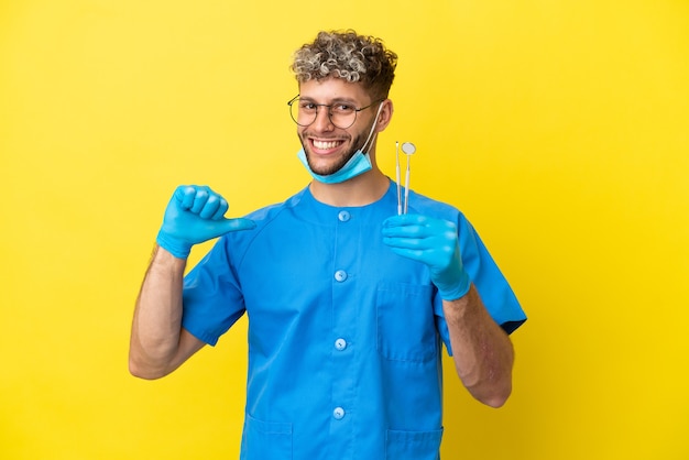 Dentiste homme caucasien tenant des outils isolés sur fond jaune fier et satisfait de lui-même