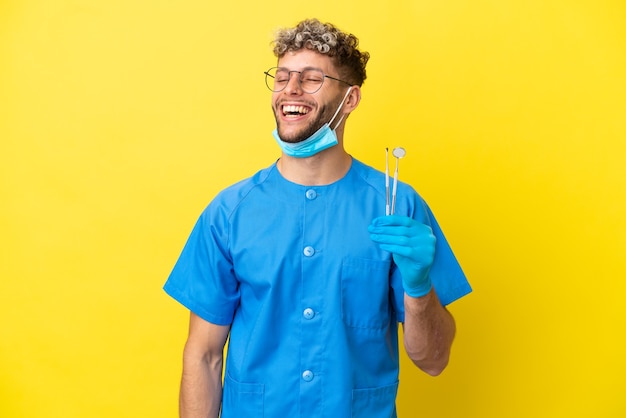 Dentiste homme caucasien tenant des outils isolés sur fond jaune en riant