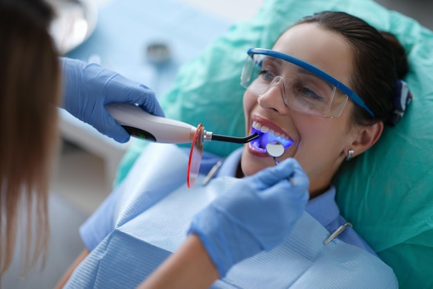 Dentiste de jeune femme dans des gants de protection vérifiant le joint dentaire fixant la lampe de photopolymère pendant le stomat...