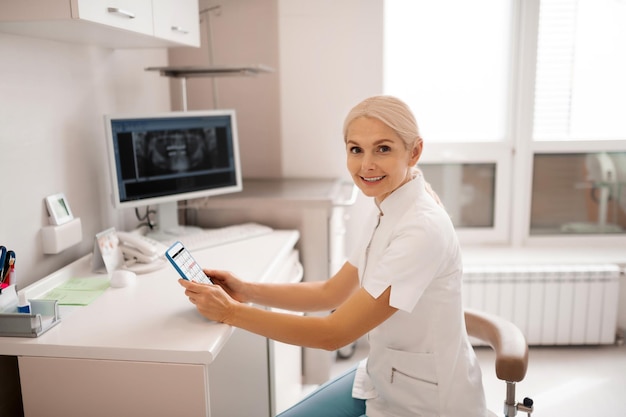 Dentiste joyeux assis à la table avec un calendrier ouvert souriant à son client entrant dans le bureau