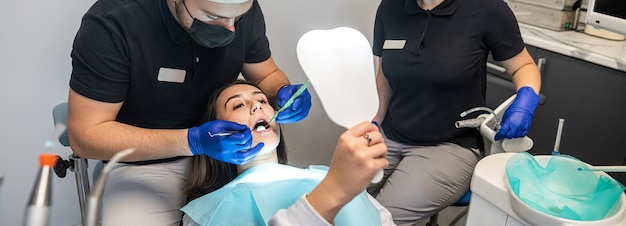 Un dentiste masculin opère une femme souriante dans une clinique dentaire et une assistante l'aide