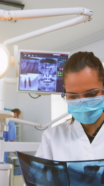 Dentiste personnalisant la lumière au-dessus de la chaise et analysant la radiographie des dents avant la chirurgie. Médecin dentiste allumant la lampe et examinant une personne portant un masque de protection et des lunettes, une infirmière l'aidant