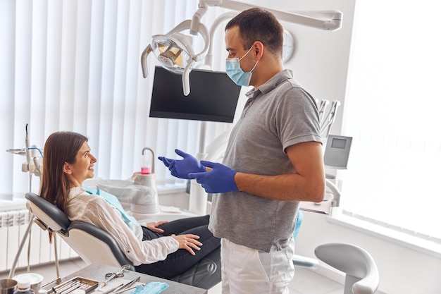 Dentiste positif joyeux et client en dentisterie. Ils se regardent et sourient. Une cliente s'assoit sur une chaise.