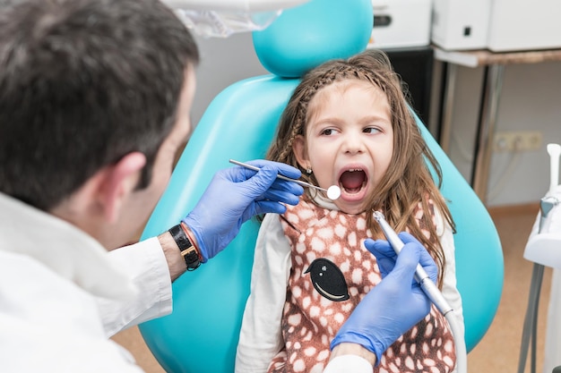 Dentiste prêt à percer une dent pour une petite fille assise dans un fauteuil dentaire avec la bouche ouverte