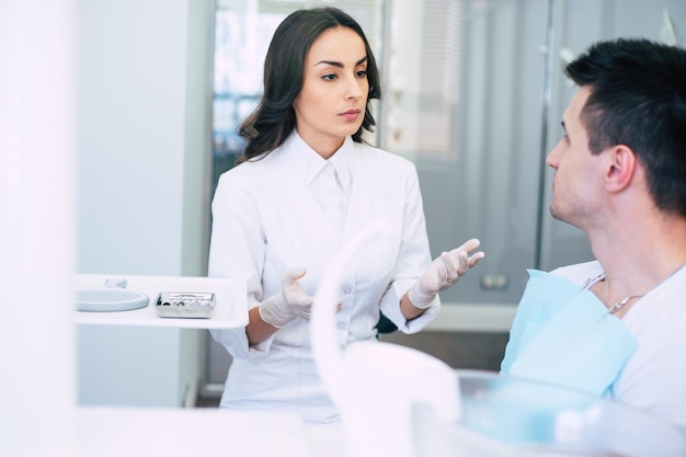 Un dentiste professionnel dans une salle de dentiste spacieuse porte des vêtements médicaux et donne quelques recommandations à son patient après son rendez-vous