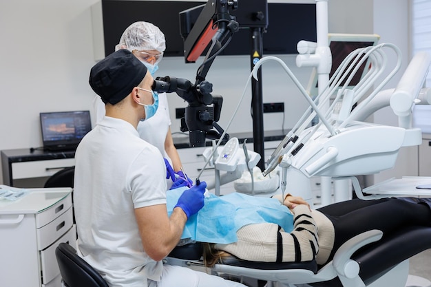 Un dentiste qui réussit regarde les dents du patient avec un microscope dentaire et tient des instruments dentaires près de sa bouche L'assistant aide le médecin Ils portent des uniformes blancs avec des masques et des gants