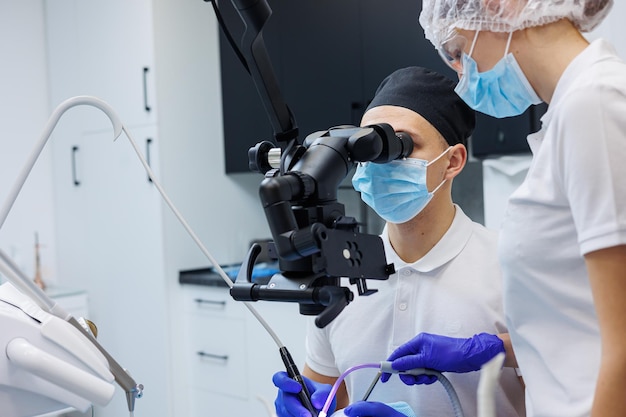 Un dentiste qui réussit regarde les dents du patient avec un microscope dentaire et tient des instruments dentaires près de sa bouche L'assistant aide le médecin Ils portent des uniformes blancs avec des masques et des gants