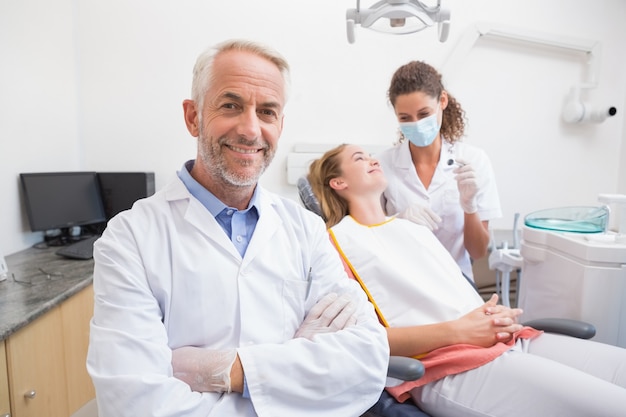 Dentiste souriant à la caméra avec assistant et patient derrière