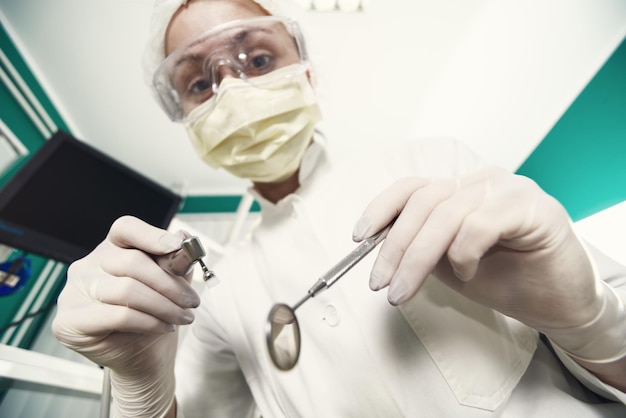 Dentiste tenant une perceuse dentaire et un miroir incliné pour inspecter un patient. Mise au point sélective.