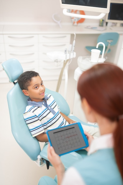 Dentiste tenant une tablette. Beau garçon mignon à la peau foncée écoutant le dentiste tenant la tablette