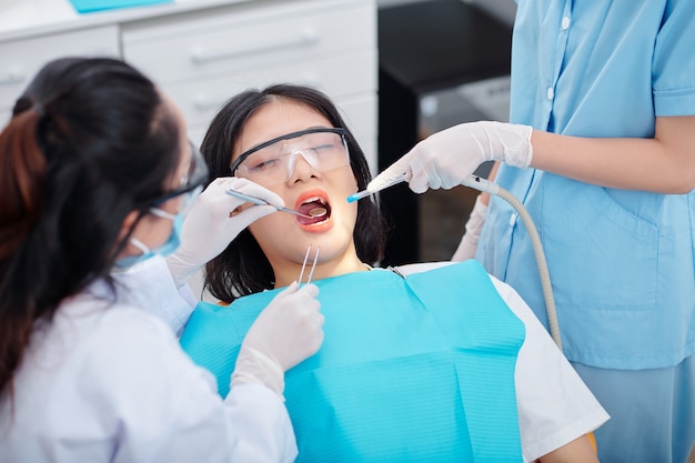 Dentiste utilisant un petit miroir lors de la vérification des dents du patient lorsque l'infirmière se tient à proximité avec un appareil d'aspiration dentaire