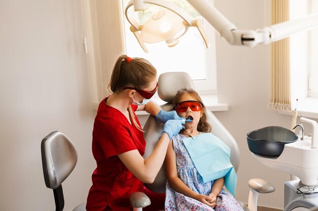 Dentisterie pour enfants Éclairage Uv de la procédure d'obturation des dents par photopolymère Enfant dentiste dans des lunettes de protection rouges traite et élimine les caries chez un patient