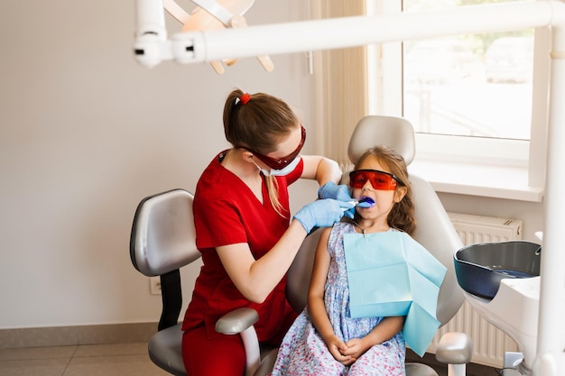 Dentisterie pour enfants Éclairage Uv de la procédure d'obturation des dents par photopolymère Enfant dentiste dans des lunettes de protection rouges traite et élimine les caries chez un patient